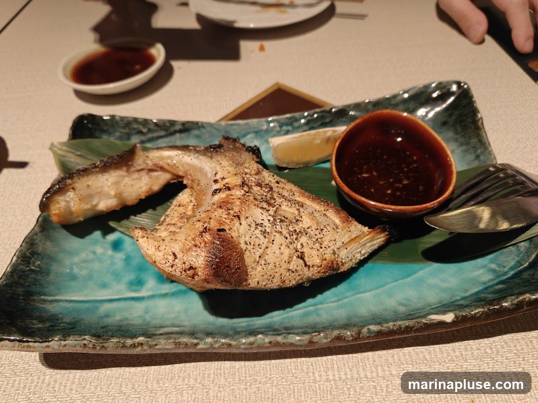 A close-up shot of a grilled fish steak served on a turquoise ceramic plate with a green leaf garnish. A small bowl of dark dipping sauce and a lemon wedge are also on the plate. Cutlery is visible to the right. Another small dish with sauce is in the background.