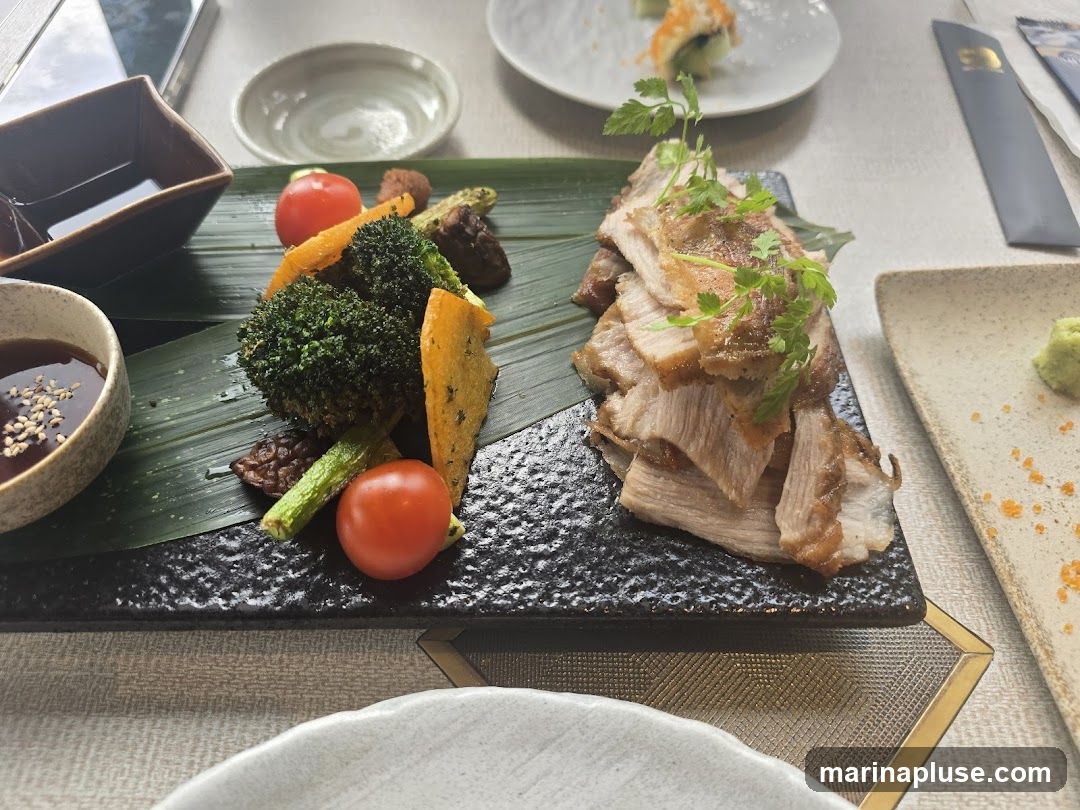A plate of sliced, roasted pork is served with a side of roasted vegetables including broccoli, carrots, cherry tomatoes, and zucchini. A small dish of dipping sauce with sesame seeds is also visible, along with a dollop of wasabi on another plate.