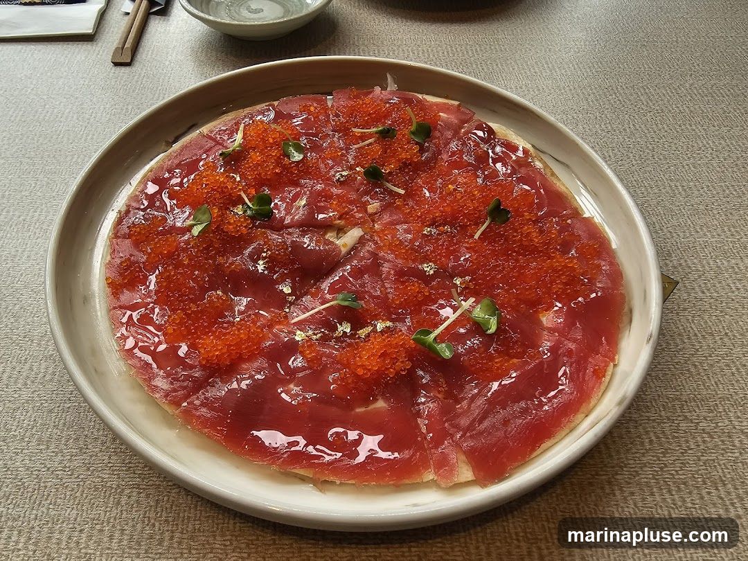 A close-up shot of a dish featuring thinly sliced raw tuna arranged on a circular base, topped with bright red roe and garnished with microgreens and edible gold flakes. The dish is served on a light-colored plate with a textured rim, placed on a neutral-toned tablecloth.