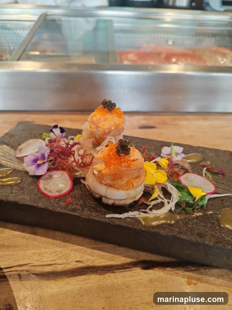 A close-up shot of a scallop appetizer served on a dark stone platter. The appetizer features two scallops, each topped with roe and black caviar. It is garnished with thinly sliced radishes, edible flowers, and shredded vegetables, with a drizzle of sauce on the platter. In the background, a display case with seafood is visible.