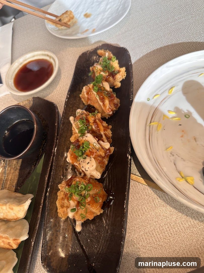 A close-up view of a meal featuring several fried appetizers drizzled with sauce and green onions, served on a dark rectangular plate. To the left, a plate of dumplings is visible. A small dish of dipping sauce is also present. Another plate with some yellow flower petals is on the right.