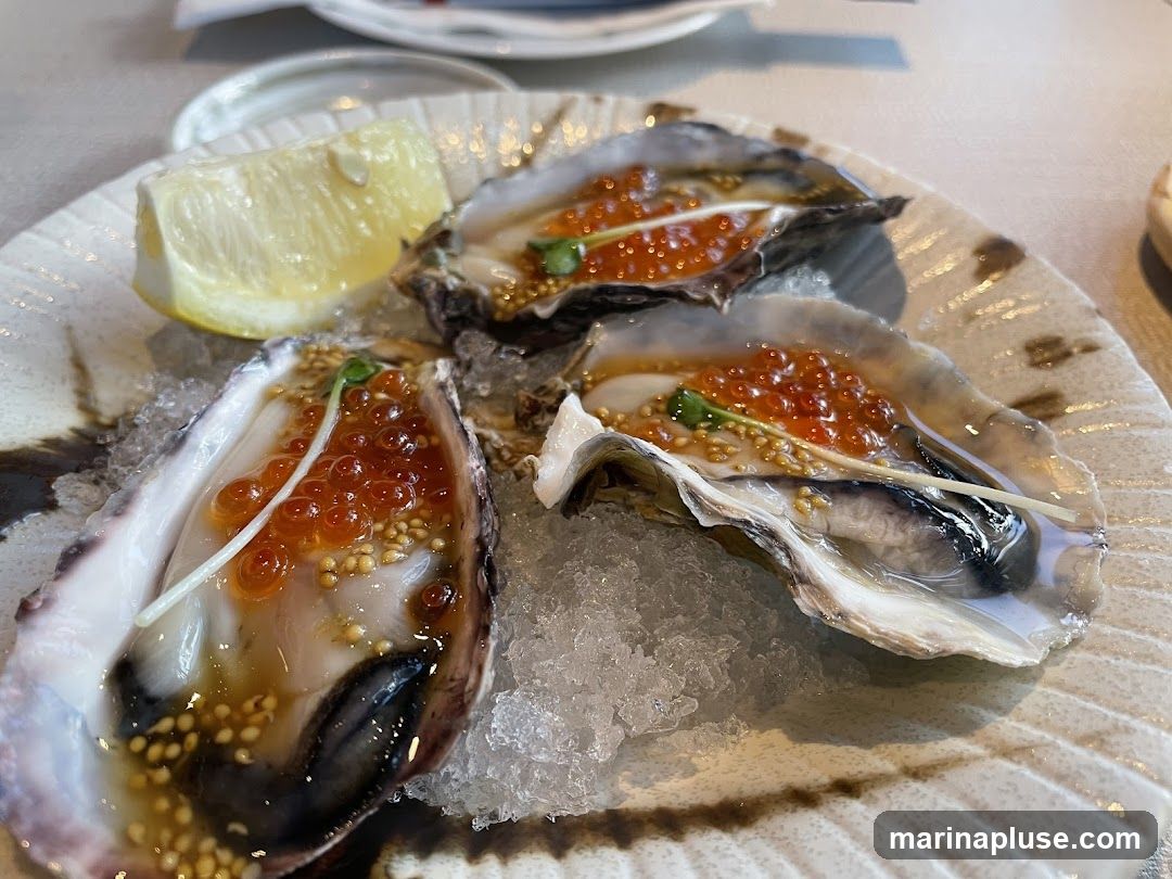 Three oysters on the half shell served on a bed of crushed ice. Each oyster is topped with ikura (salmon roe), a light sauce, and a garnish of microgreens. A wedge of lemon is placed on the side of the plate. The plate has a textured, speckled pattern.