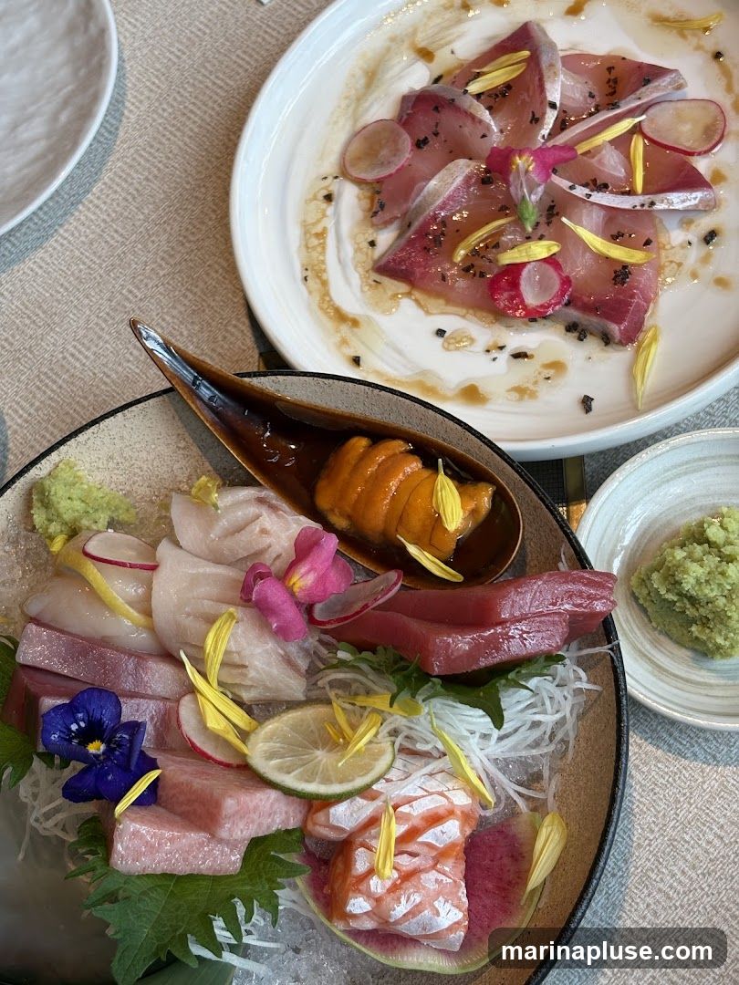 A close-up overhead view of a sashimi platter featuring various types of raw fish, sea urchin, and garnishes. The platter is arranged on a bed of shredded daikon radish and ice, with edible flowers and thinly sliced radishes. A small dish of wasabi is visible to the right. Another plate with thinly sliced fish, radishes, and a light dressing is in the background.