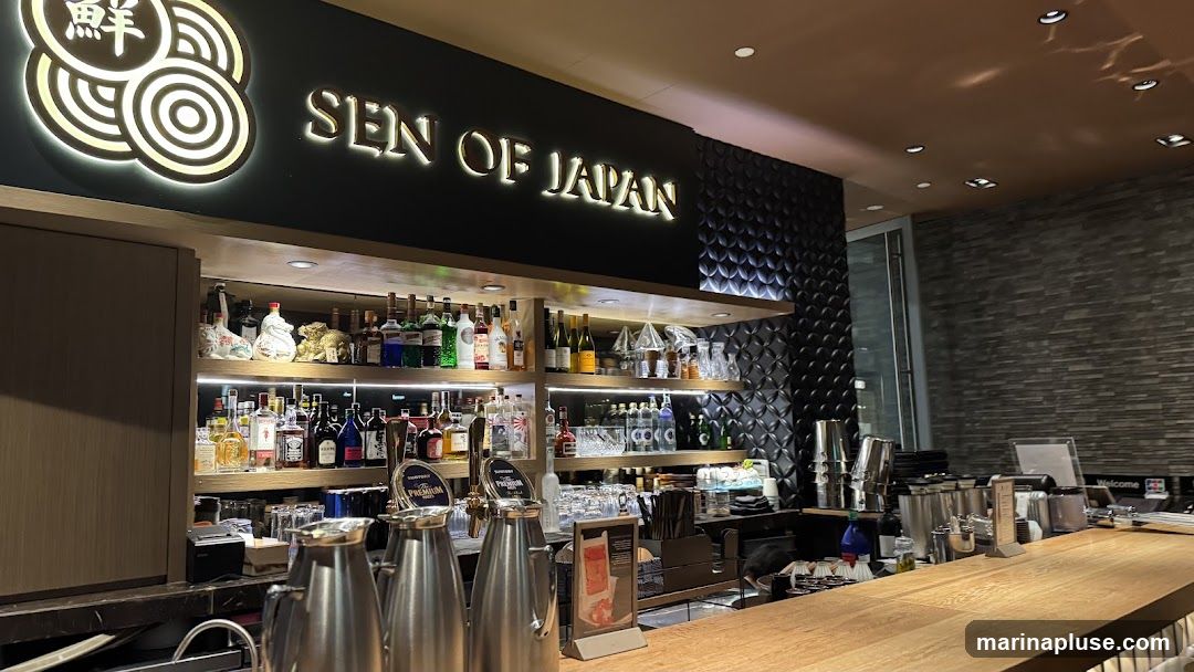 The image shows the interior of a bar with a prominent sign that reads "SEN OF JAPAN" and a circular logo with Japanese characters. Shelves behind the bar are stocked with various bottles of liquor and other beverages, as well as glassware. The bar itself is made of wood, and there are metal beverage dispensers visible. The background features textured dark walls.