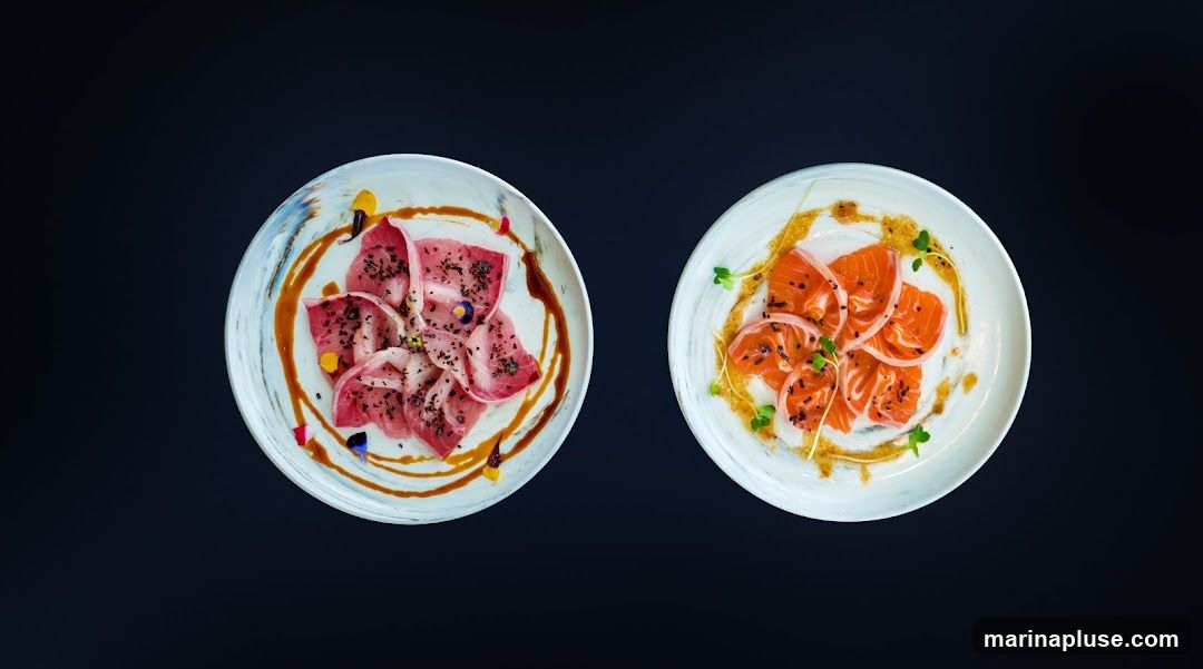 Two plates of thinly sliced raw fish, garnished with herbs and sauce, are presented against a dark background. The plate on the left features pinkish-red slices, possibly tuna or hamachi, sprinkled with black sesame seeds and edible flowers. The plate on the right displays vibrant orange salmon slices arranged with thinly sliced onions and microgreens. Both dishes are drizzled with a light-colored sauce and a darker, savory sauce.