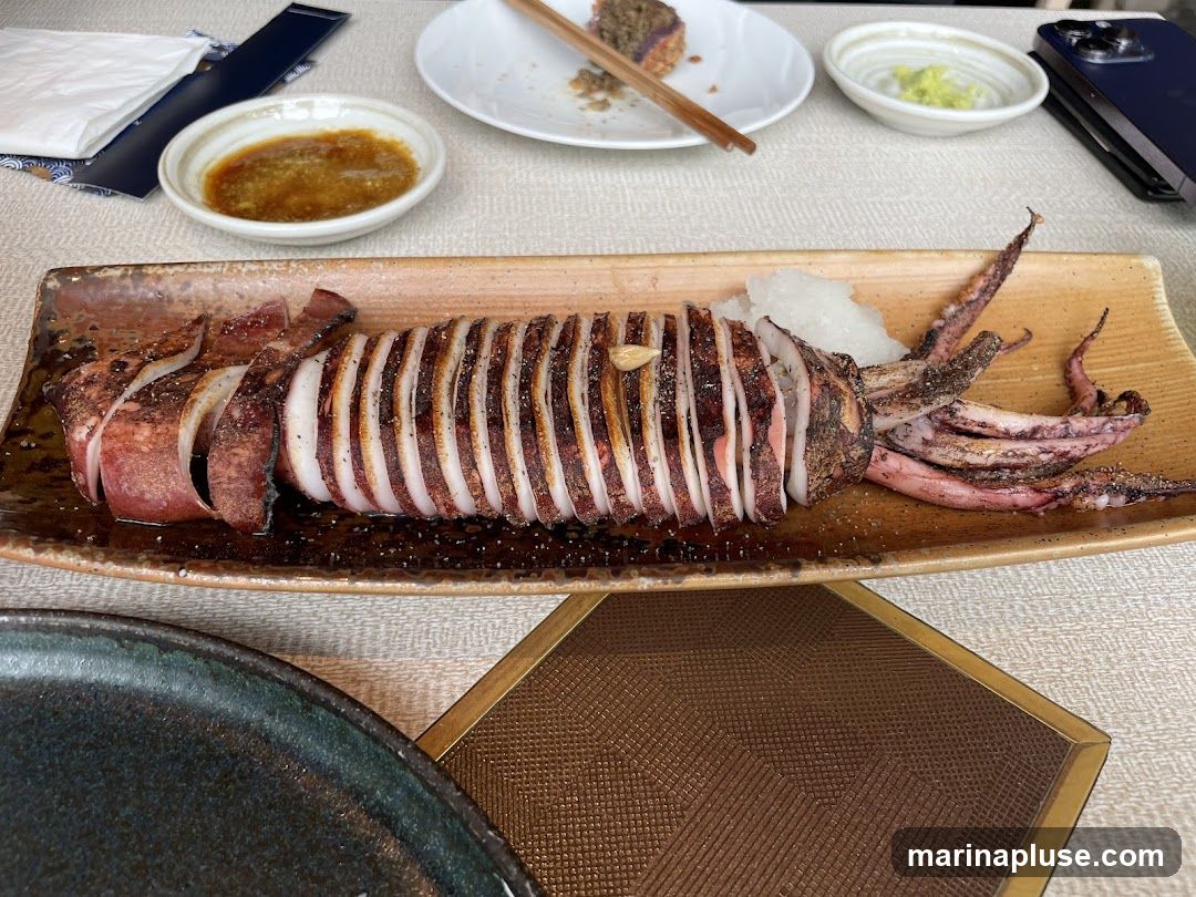 A whole grilled squid, sliced and served on a rectangular platter with grated daikon radish and a dipping sauce. The squid is seasoned with salt and pepper. In the background, there are small bowls of sauce and what appears to be a piece of cake.
