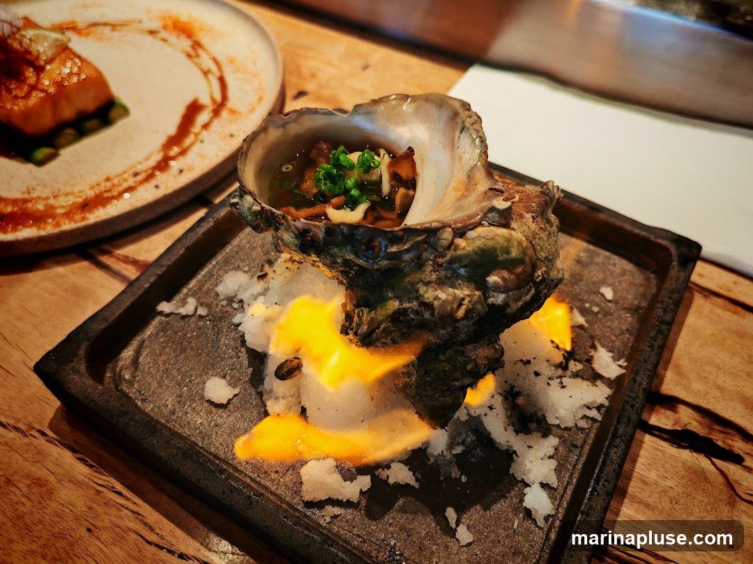 A close-up shot of a single oyster served in its shell, placed on a bed of salt and lit with a flame. The oyster contains a small portion of broth and is garnished with chopped green onions and other ingredients. In the background, a plate with a piece of salmon and asparagus is visible. The dishes are set on a wooden table.