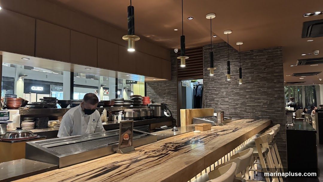 A chef in a white uniform and mask works behind a sushi bar. The bar has a live-edge wooden countertop with several stools. Behind the chef, kitchen equipment and stacks of plates are visible. The background shows a reflection of a shopping mall with a "CLUB 21" sign.