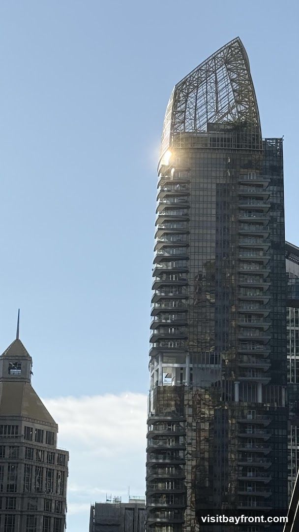 A tall, modern skyscraper with a distinctive, open-lattice crown structure dominates the frame against a clear blue sky. The building features a facade of dark glass and numerous balconies that jut out at various levels. Sunlight glints off the upper portion of the skyscraper, highlighting its architectural details. To the left, another building with a more traditional, stepped roofline and visible construction elements is partially visible.
