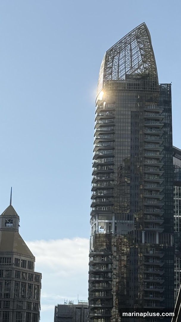 A tall, modern skyscraper with a distinctive, open-lattice crown structure dominates the frame against a clear blue sky. The building features a facade of dark glass and numerous balconies that jut out at various levels. Sunlight glints off the upper portion of the skyscraper, highlighting its architectural details. To the left, another building with a more traditional, stepped roofline and visible construction elements is partially visible.