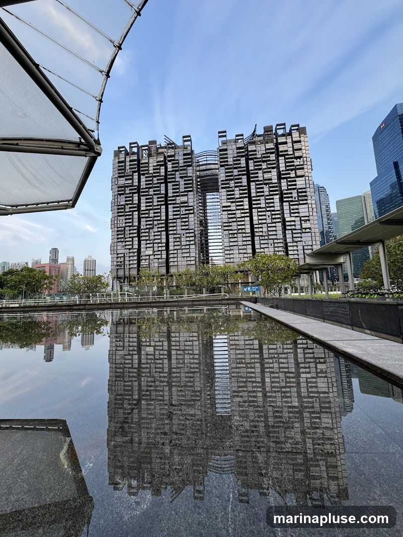 A striking modern building with a unique, modular facade is reflected in a dark, still body of water. The building's design features numerous rectangular windows and protruding elements, creating a textured and complex appearance. In the foreground, a portion of a large, white, curved canopy structure is visible, adding an architectural element to the scene. The sky above is a clear blue with faint wisps of clouds. The reflection in the water mirrors the building and the sky, creating a symmetrical and visually engaging composition. Other buildings and greenery are visible in the background, suggesting an urban environment.