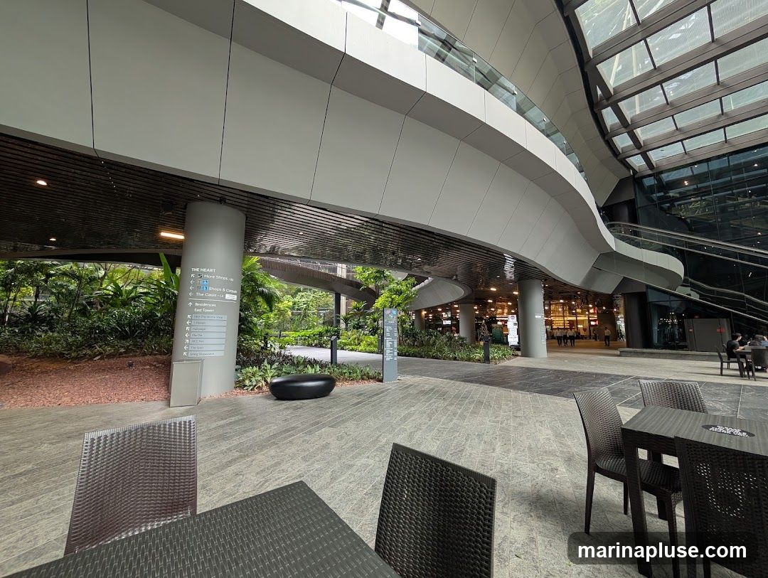 An outdoor seating area with tables and chairs is set on a paved walkway. In the background, a modern building with a curved facade and a glass roof is visible. Lush greenery surrounds the area, and a directional signpost indicates various locations within "The HEART." The scene suggests a commercial or public space with a focus on contemporary design and nature integration.
