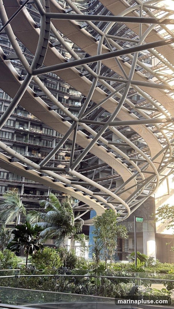 A view looking up at a complex, modern architectural structure with a metallic framework and woven walkways. Below the structure, a vibrant garden with various tropical plants and palm trees is visible. The background shows a multi-story building with many windows, reflecting the sunlight. An illuminated green 'EXIT' sign is mounted on a pole to the right.