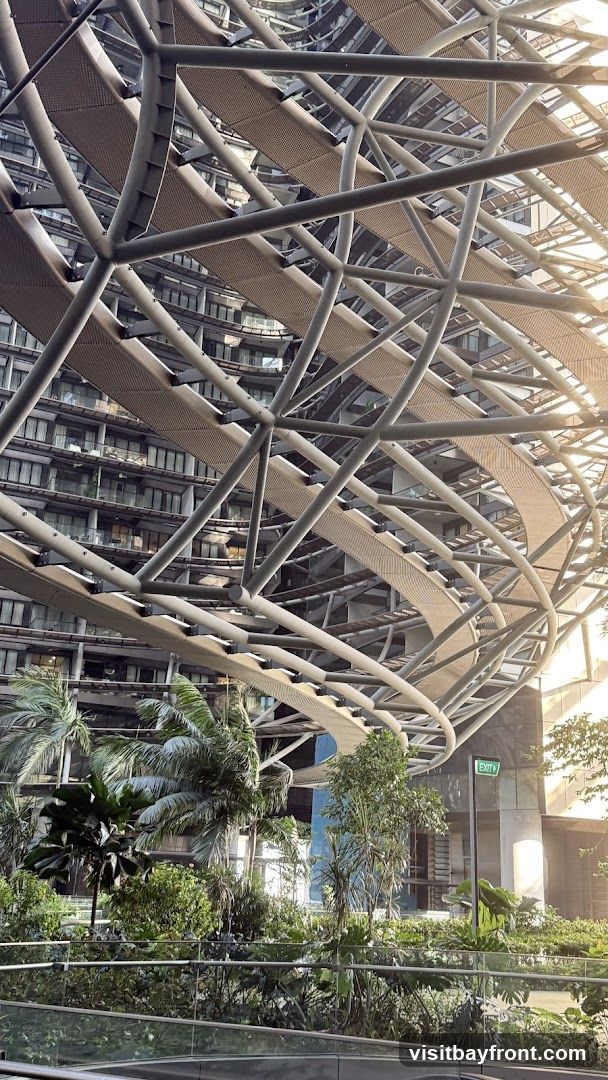 A view looking up at a complex, modern architectural structure with a metallic framework and woven walkways. Below the structure, a vibrant garden with various tropical plants and palm trees is visible. The background shows a multi-story building with many windows, reflecting the sunlight. An illuminated green 'EXIT' sign is mounted on a pole to the right.