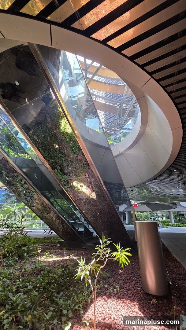 An interior view of a modern building with a striking architectural design. A large, curved structure with reflective surfaces dominates the foreground, featuring a vertical garden with lush green plants. Sunlight streams in from a large, circular opening in the ceiling, illuminating the space and casting shadows. The ceiling itself is a complex structure of glass and metallic elements. In the lower right, a cylindrical metallic object stands amidst gravel and small plants. The overall impression is one of a contemporary, nature-integrated urban space.