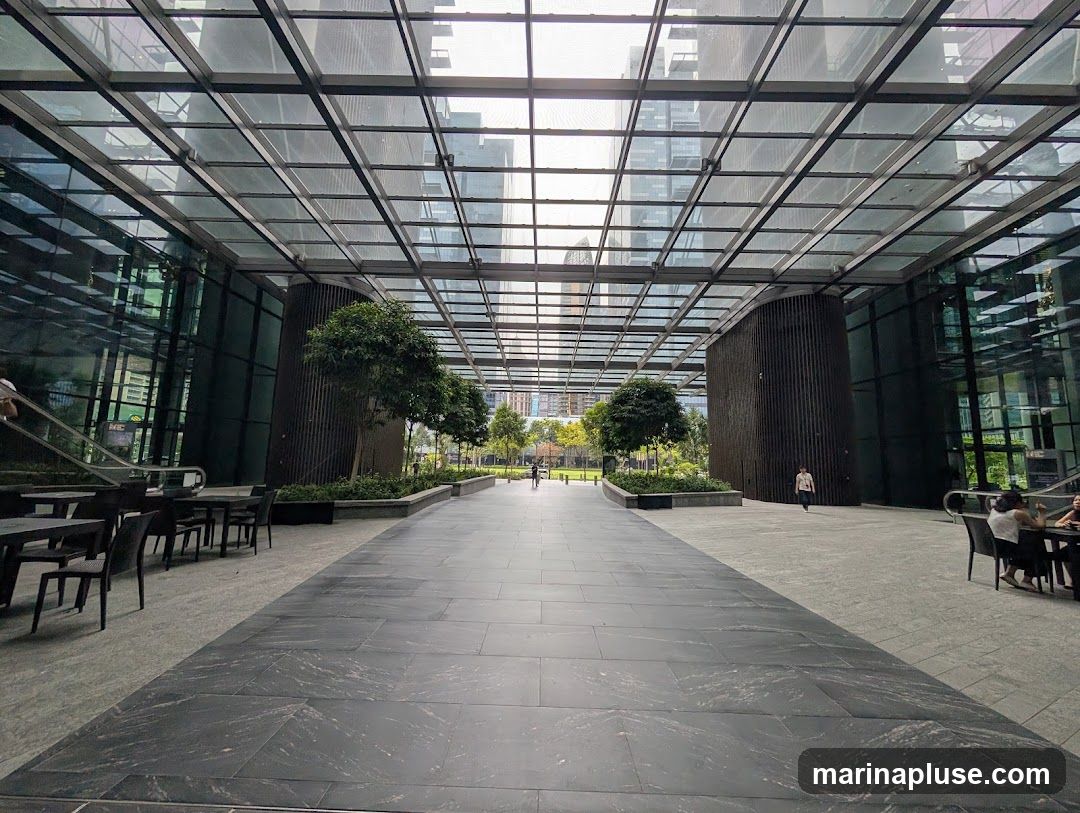 An expansive, modern outdoor seating area with a glass ceiling, flanked by tall buildings. The walkway is paved with dark, polished stone, leading towards a green space with trees. Tables and chairs are set up on either side, with a few people seated and walking in the distance.