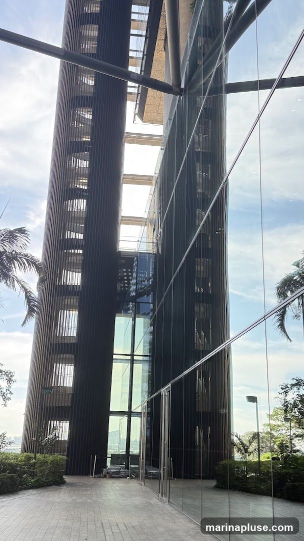 A view of a modern building with a distinctive curved facade made of dark vertical slats. Adjacent to it is a glass-fronted section reflecting the sky and surrounding greenery. The walkway in front is paved with stone tiles. An 'EXIT' sign is visible on the slatted section of the building.