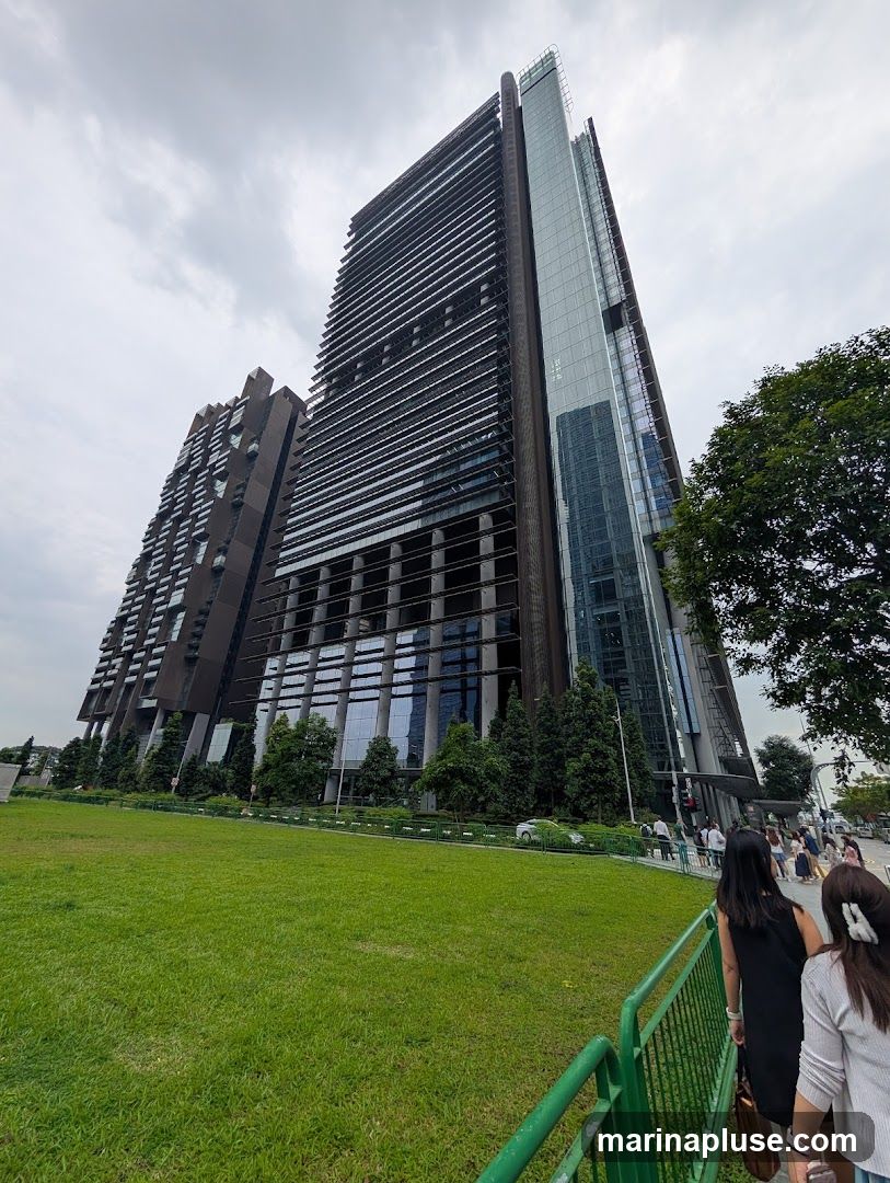 A wide-angle shot captures the imposing modern architecture of two skyscrapers, one with a dark, slatted facade and the other with a sleek glass exterior. They are set against a cloudy sky. In the foreground, a lush green lawn slopes gently, bordered by a dark green fence. Beyond the lawn, a tree-lined pathway leads to the entrance of the buildings, where several people are walking. A white car is visible on the road in the distance.