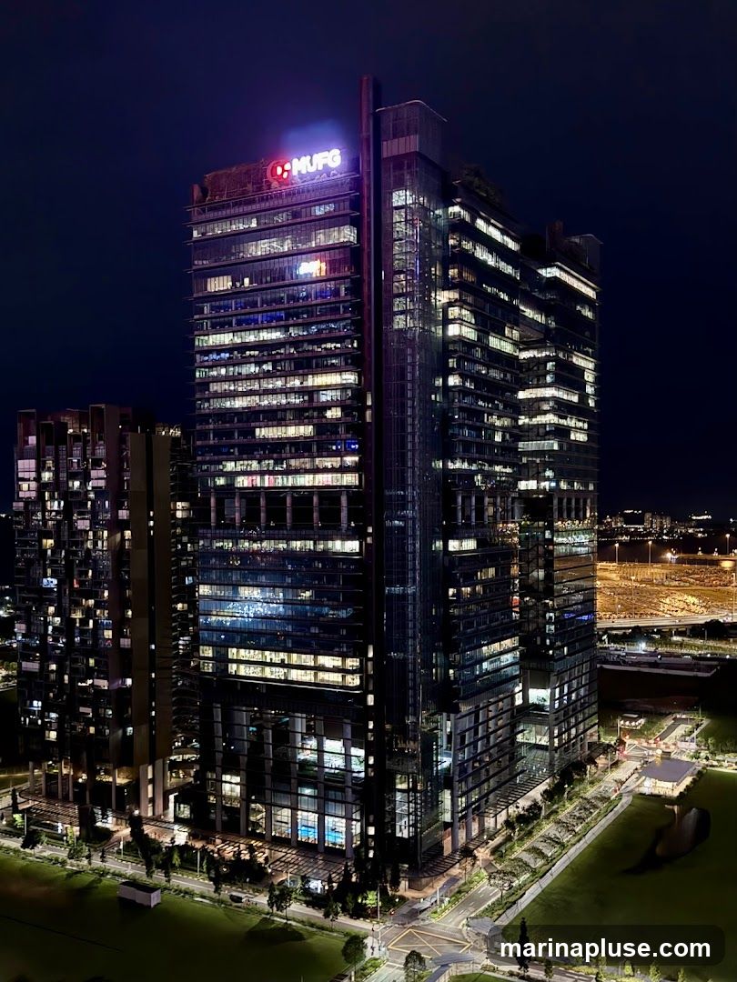A nighttime aerial view of a modern skyscraper with the MUFG logo illuminated at the top. The building is surrounded by other structures and green spaces, with lights visible in many of the windows, suggesting occupancy.
