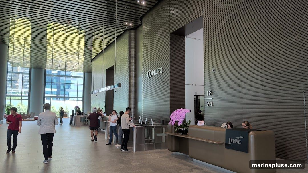 A spacious and modern office lobby with a high ceiling featuring a reflective, slatted design. Large windows offer a view of city buildings. Several people are seen walking through the lobby, and some are interacting at a reception desk. The MUFG logo is prominently displayed on a wall, and the PwC logo is visible on the reception desk. Elevator numbers 16 and 24 are also indicated. A large floral arrangement adds a touch of color to the scene.