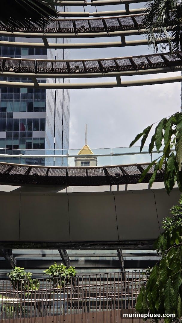 A view through a modern architectural structure, possibly a skywalk or elevated walkway, revealing a cityscape. In the background, a tall building with a distinctive golden spire stands out against a cloudy sky. The foreground features curved walkways with decorative metal grating and lush green foliage. The overall scene showcases a blend of contemporary urban design and natural elements.