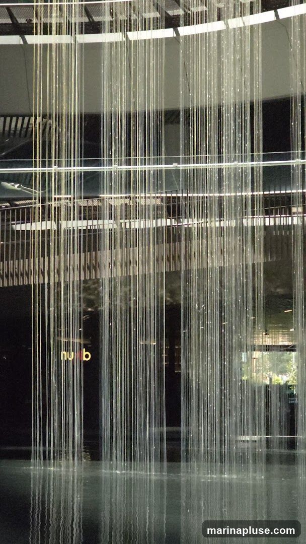 A water feature with numerous thin, illuminated strands cascading into a pool of water. Behind the water feature, a dark facade is visible with a glowing sign that reads "nub". The overall impression is modern and artistic.