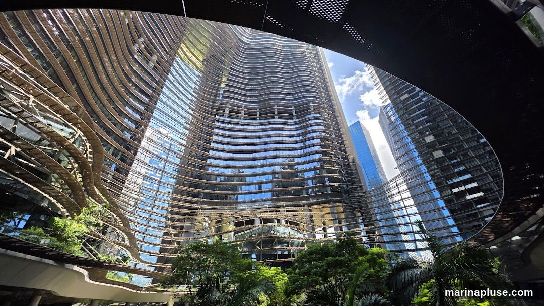 A wide-angle shot looking up at a modern skyscraper with a unique, wavy facade. The building is covered in glass and horizontal wooden or metallic slats that create a textured, organic appearance. Lush green trees and foliage are visible at the base and on balconies, contrasting with the sleek urban structure. The sky above is blue with scattered clouds.
