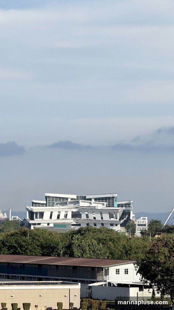 A wide shot captures a striking modern building with a complex, geometric facade, set against a backdrop of trees and a hazy sky. The building features a white exterior with numerous angled white panels and glass windows, creating a dynamic and layered appearance. In the foreground, there are lower structures with corrugated roofs and some smaller buildings. The overall scene suggests an urban or industrial landscape with contemporary architecture.