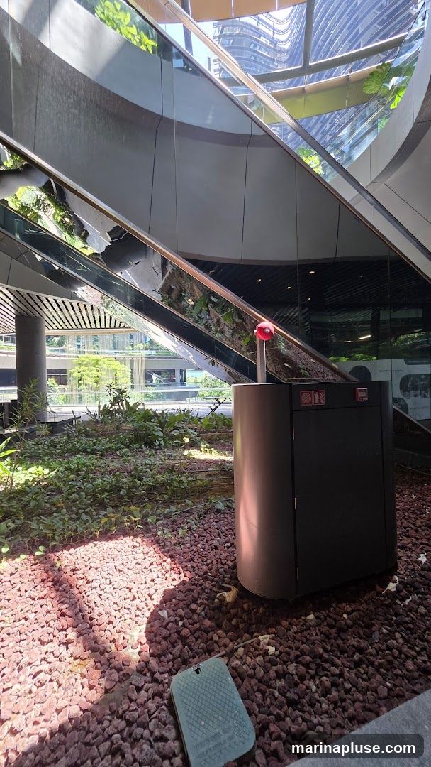An interior view of a modern building atrium featuring a curved escalator with reflective surfaces. Lush green plants and small trees are planted in a bed of red gravel surrounding the base of the escalator. A dark grey cylindrical structure with a red light on top stands in the foreground, and a green utility box is visible on the ground. The background shows the exterior of other buildings through large glass windows.