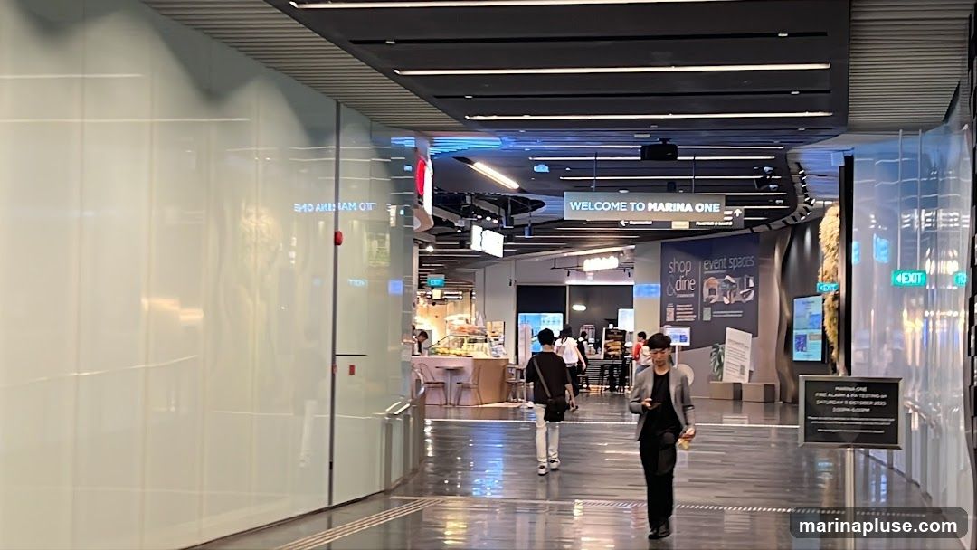 A wide, well-lit corridor in a modern shopping mall. On the left, a large, frosted glass wall reflects the interior. On the right, there are various signs and displays. One sign reads "WELCOME TO MARINA ONE" with directional arrows. Another sign indicates "shop & dine" and "event spaces". Further down, an "EXIT" sign is visible. People are walking through the corridor, and some are browsing at a food stall with display cases filled with food items. The floor is highly reflective, mirroring the ceiling lights and architectural features.