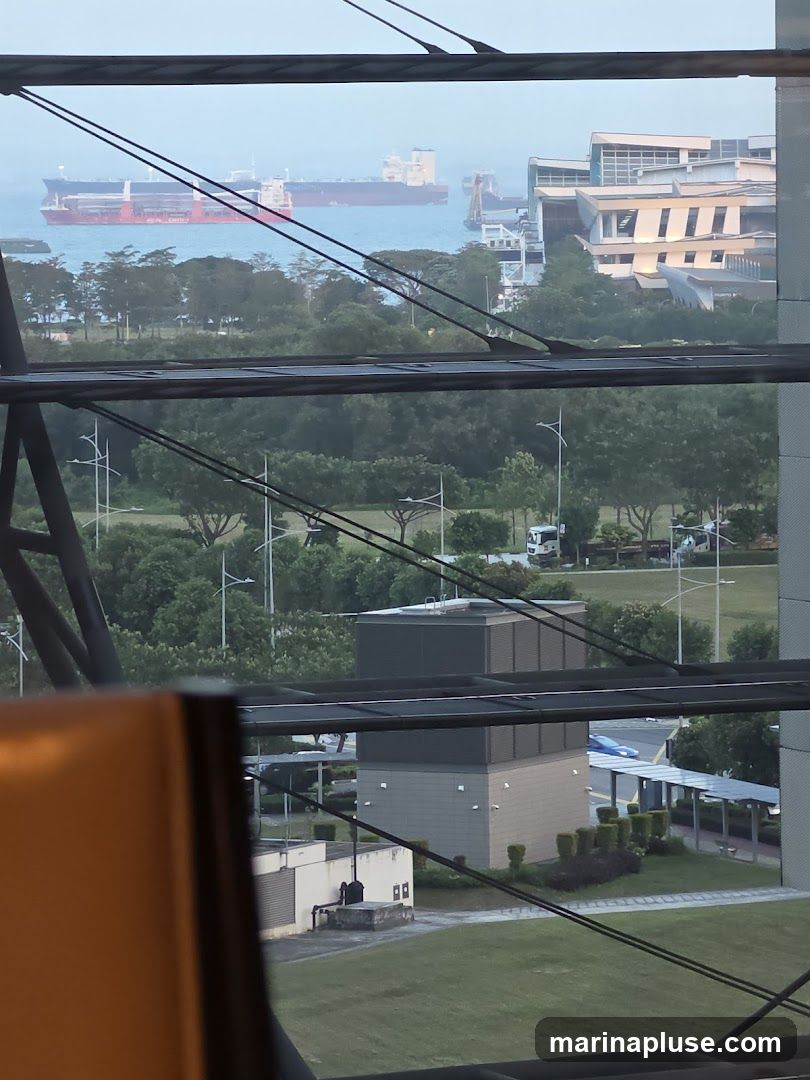 A view from a window overlooking a body of water with several large ships docked or sailing. In the middle ground, there are green trees and some industrial-looking buildings. The foreground shows a portion of an orange chair and structural elements of the building the photo was taken from.