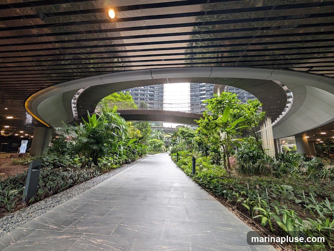 A paved walkway curves through a landscaped area with abundant green plants and trees. Overhead, a modern architectural structure with a reflective ceiling and curved elements creates a covered walkway or bridge. In the background, tall buildings are visible. The scene suggests a contemporary urban environment with a focus on integrating nature.