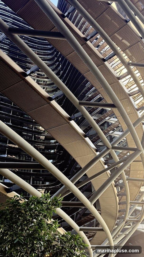 A low-angle shot captures a modern architectural design featuring a curved walkway with a metal grate surface supported by a complex network of cylindrical metal beams. The walkway spirals upwards, creating a dynamic visual effect. In the background, a building with a dark, curved facade and numerous windows is visible. A lush green tree with small white flowers is partially in the foreground, adding a touch of nature to the urban structure. The lighting suggests it is daytime, with sunlight casting shadows and highlighting the metallic elements.