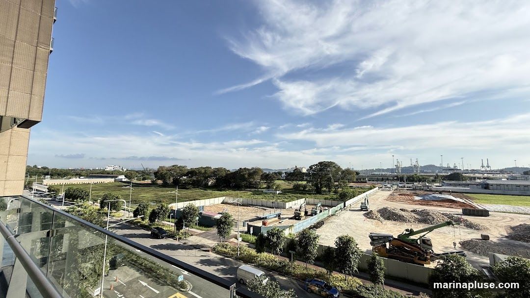 A wide shot of a construction site under a bright blue sky with scattered clouds. In the foreground, a modern building's facade and a glass balcony railing are visible on the left. The construction area features various heavy machinery, including excavators and a large crane, along with piles of dirt and debris. In the background, a port with numerous cranes and industrial structures stretches across the horizon. The scene suggests ongoing development and industrial activity.