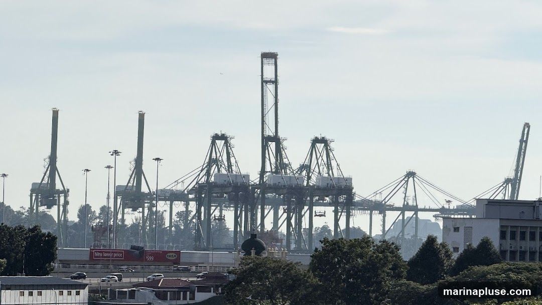 A wide shot of a port with numerous large cranes silhouetted against a hazy sky. In the foreground, a train car with an advertisement for IKEA is visible, featuring the text "New lower price" and "$599". Trees and buildings are also present in the lower part of the image.