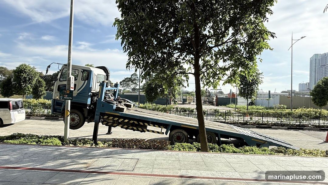 A flatbed truck is parked on a sidewalk, with its ramp extended. The truck is blue and white. In the background, there are trees, construction fencing, and tall buildings. A white van is parked to the left of the truck.