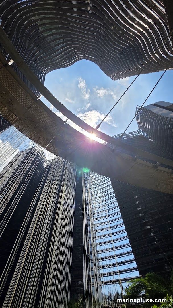 A low-angle shot looking up at modern buildings with curved facades. A sun flare is visible in the sky, with clouds. Hanging strings or cables create a curtain-like effect in the foreground.
