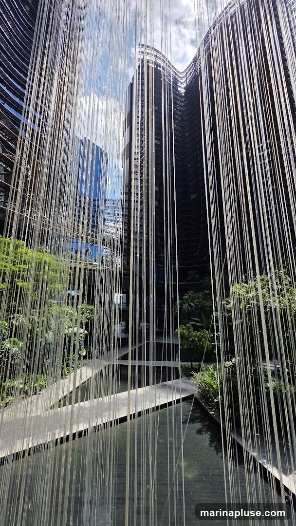 A view through a curtain of thin, light-colored strands, revealing a modern courtyard. The strands hang from above, creating a textured foreground. Beyond them, contemporary buildings with curved facades rise against a blue sky with scattered clouds. The courtyard features pathways over dark water features and lush green foliage, suggesting a tranquil, urban oasis.