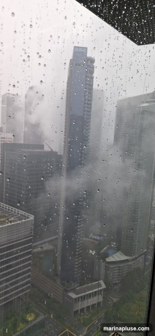 A view of a city skyline through a window covered in raindrops. Tall buildings are visible, some shrouded in mist or clouds. The weather appears overcast and rainy.