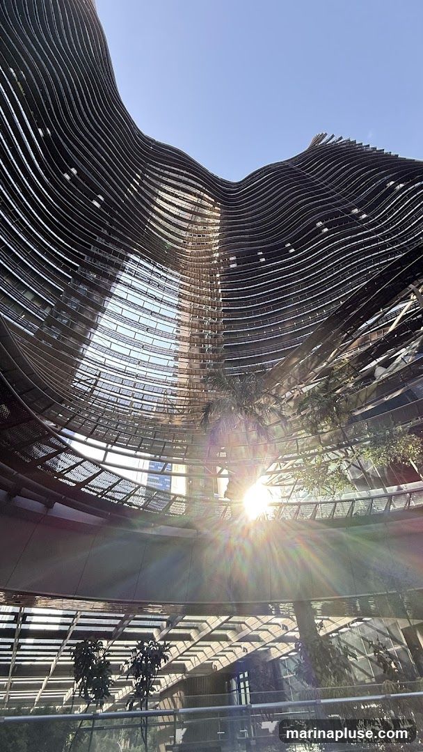 A low-angle shot looking up into the interior of a modern building with a curved, wave-like facade. The structure is made of dark, horizontal slats that create a rhythmic pattern. Sunlight streams in from the top, creating a lens flare effect and casting shadows on the interior levels. Trees and greenery are visible on some of the interior balconies, adding a touch of nature to the architectural design. The overall impression is one of contemporary design and natural light integration.