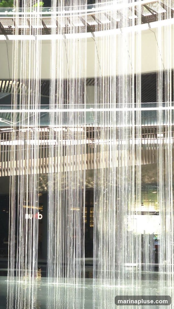 A contemporary building facade is partially obscured by a curtain of thin, vertical strands, possibly part of a water feature or decorative installation. The strands create a shimmering effect as they descend into a pool of water at the base, reflecting the light and the building's architecture. Behind the strands, the building features multiple levels with glass windows and architectural elements, suggesting a commercial or public space. A partial logo or sign, appearing as 'nu b', is visible on the glass in the lower left portion of the image.