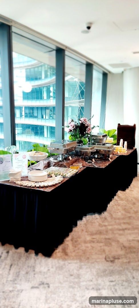 A long table covered with a black tablecloth is set up with a buffet. There are several chafing dishes, bowls of food, and platters of appetizers. A floral arrangement is placed in the center of the table. In the background, large windows offer a view of modern buildings. Disposable plates and cutlery are stacked on the left side of the table.