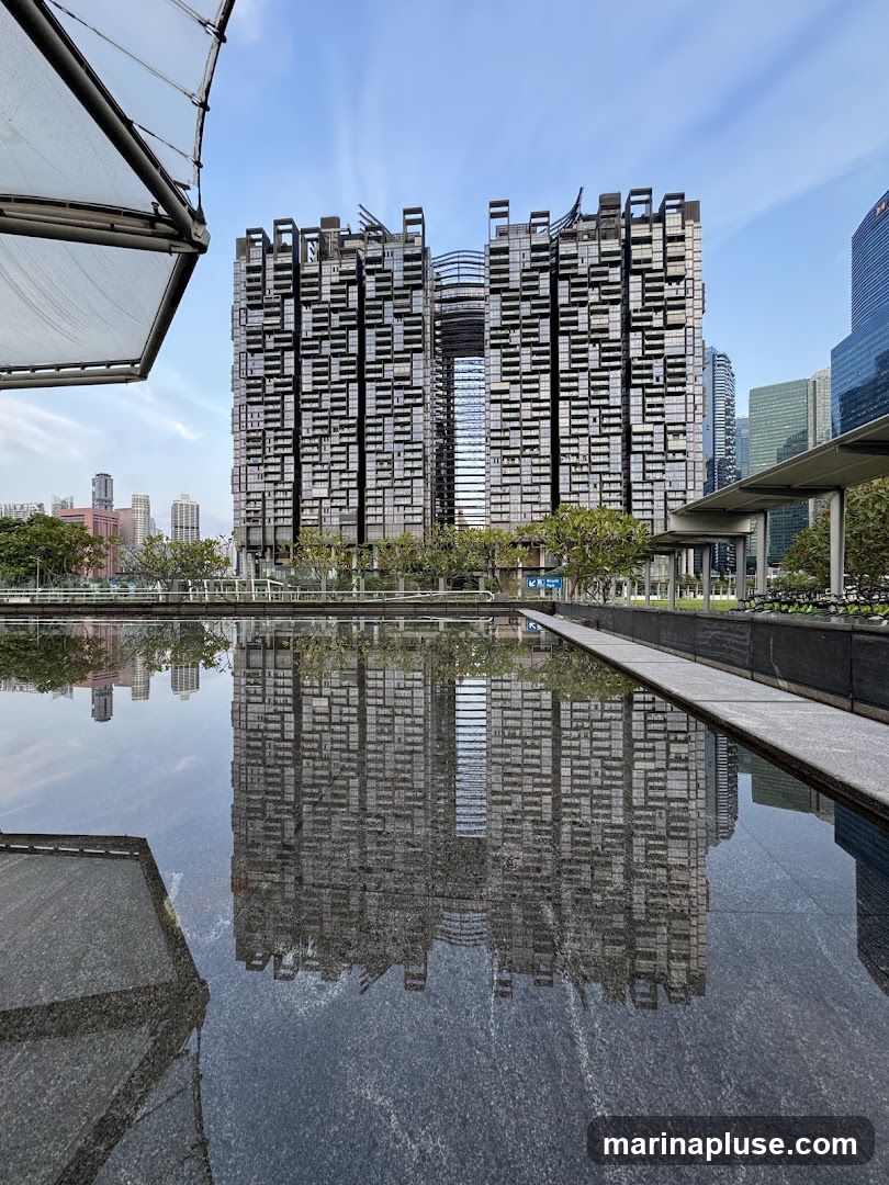 A striking modern building with a unique, modular facade is reflected in a calm body of water. The building features a complex arrangement of dark rectangular units, creating a textured and visually dynamic appearance. A central opening or gap breaks the symmetry of the structure. The sky above is a clear blue with wispy clouds. In the foreground, the water's surface mirrors the building and the sky, creating a symmetrical and serene composition. The surrounding area appears to be a well-maintained urban landscape with trees and other contemporary architecture visible in the distance.