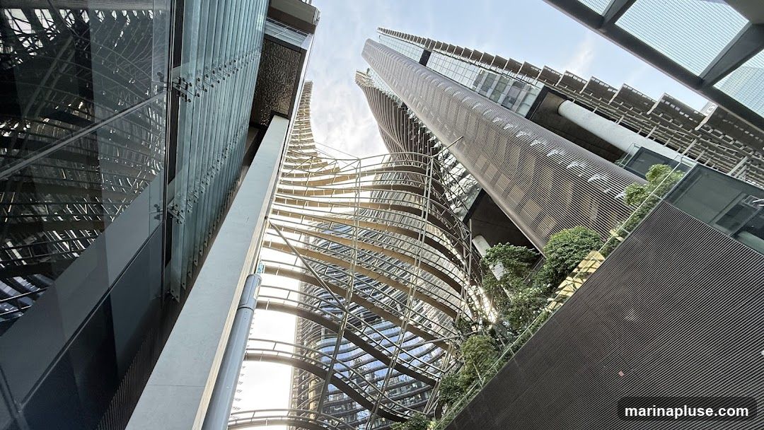 A low-angle shot looking up at several modern skyscrapers with unique architectural designs. The buildings feature a mix of glass, metal, and textured facades, with some incorporating green spaces and intricate structural elements. The sky is visible above, with a bright, slightly overcast appearance.