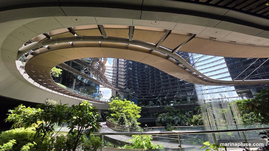 A wide-angle view of a contemporary architectural space featuring a curved, open-air structure with a woven canopy overhead. Below, multiple levels of walkways and balconies are surrounded by abundant tropical plants and trees. In the background, modern high-rise buildings with reflective glass facades rise towards a bright sky. A decorative element resembling a waterfall made of thin strands cascades down one of the levels.