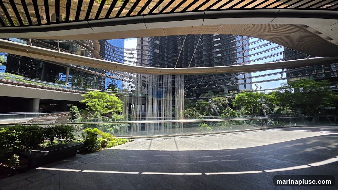A wide-angle view of a contemporary architectural space featuring a curved walkway in the foreground, leading towards a central atrium. The atrium has a striking waterfall-like feature composed of numerous vertical metallic strands. Surrounding the atrium are multi-story buildings with reflective glass facades, interspersed with abundant tropical greenery, including trees and various plants. The ceiling above is a metallic, slatted structure that reflects the light and surroundings. The overall impression is one of a modern, urban oasis that blends nature with sophisticated design.