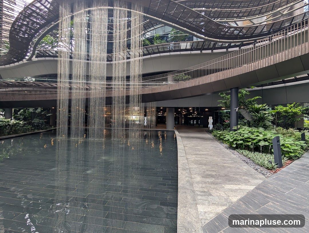 An indoor courtyard features a large pool of water with a cascading waterfall made of thin strands. A walkway with stone paving leads past lush greenery and planters. The architecture includes multiple levels with curved walkways and a metal mesh ceiling structure.