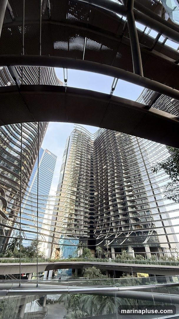 A view looking up at several modern skyscrapers with unique, wavy facades. The buildings are made of glass and metal, reflecting the sky and surrounding structures. In the foreground, there is a walkway or bridge with greenery, and the overall scene is framed by an overhead architectural structure with a perforated metal ceiling.