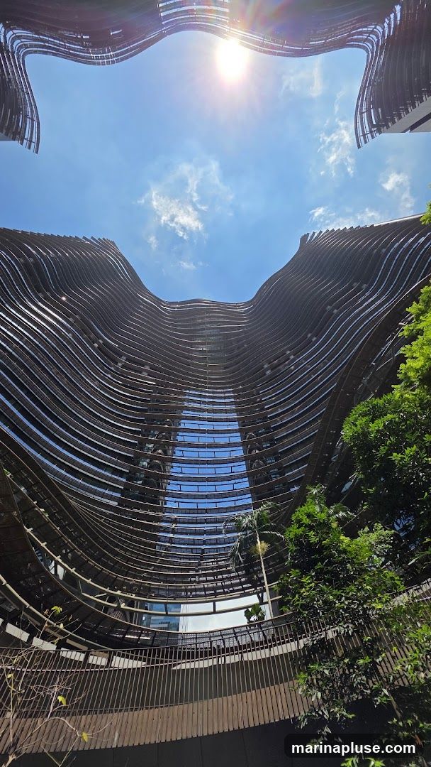 A low-angle shot looking up at a modern building with a distinctive wavy facade. The building's exterior is composed of horizontal, curved elements that create a rippling effect. Sunlight streams down from a bright blue sky with scattered clouds, reflecting off the windows of the building. Lush green trees and foliage are visible on the right side, adding a natural element to the architectural scene. A walkway with a railing is visible in the foreground.