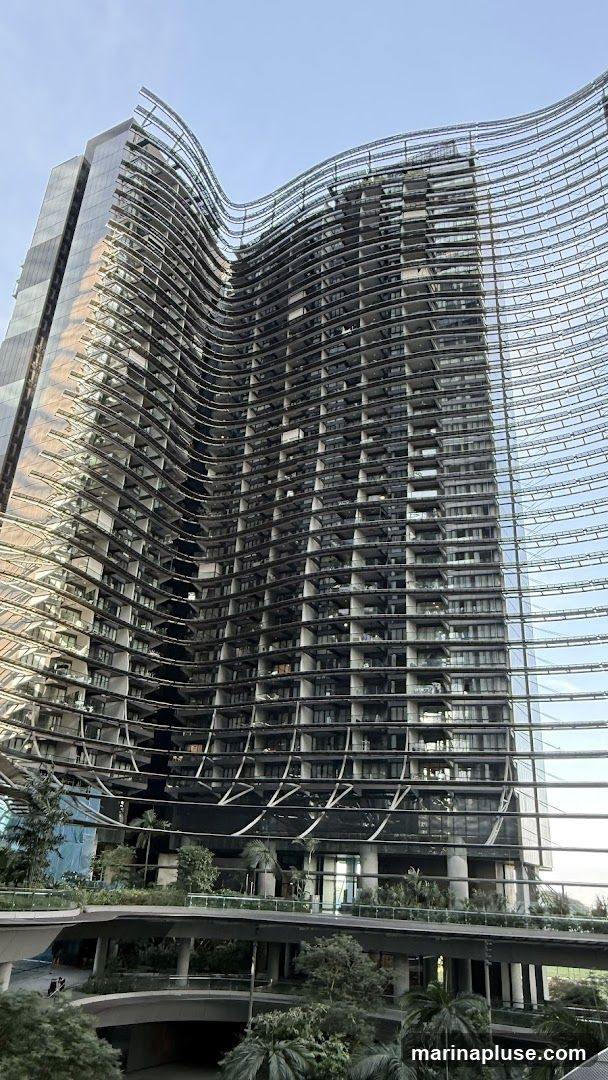 A low-angle shot of a modern skyscraper with a distinctive wavy facade. The building features a glass exterior on one side and a series of balconies or open-air spaces on the other, creating a textured, undulating appearance. Below the main structure, there are elevated walkways and lush greenery, suggesting a complex with integrated landscaping. The sky above is clear and blue.